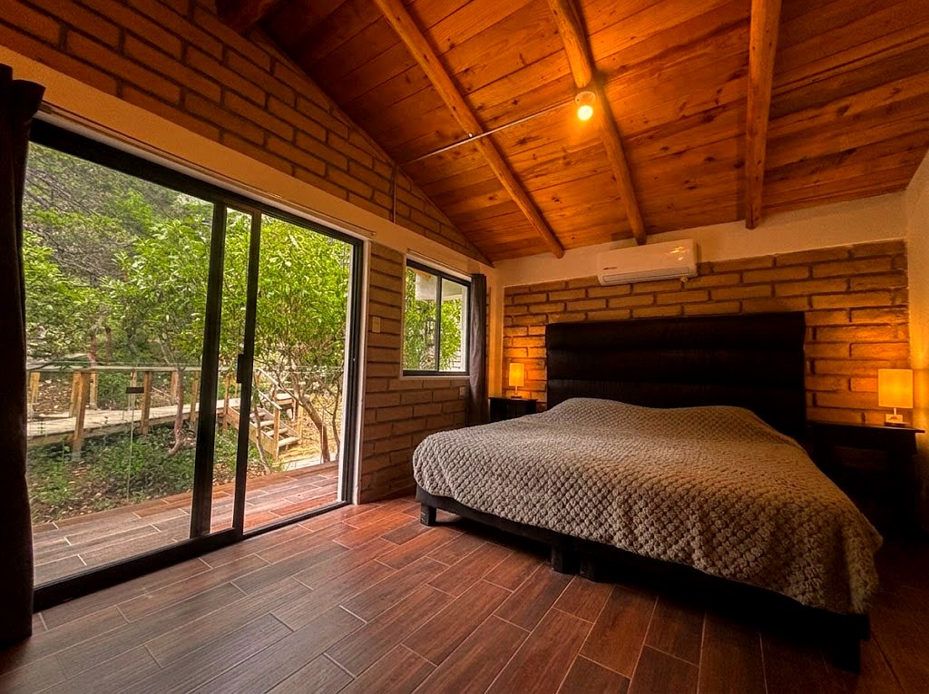 Recámara principal de planta alta en OSA MAYOR con cama king, ventanal al bosque y vigas de madera