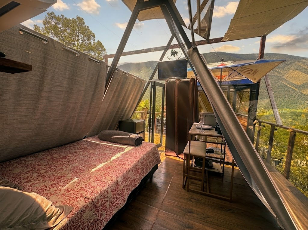 Interior de SIRIUS con cama matrimonial, cortinas texturizadas, Smart TV y ventanal A-frame con vista al bosque