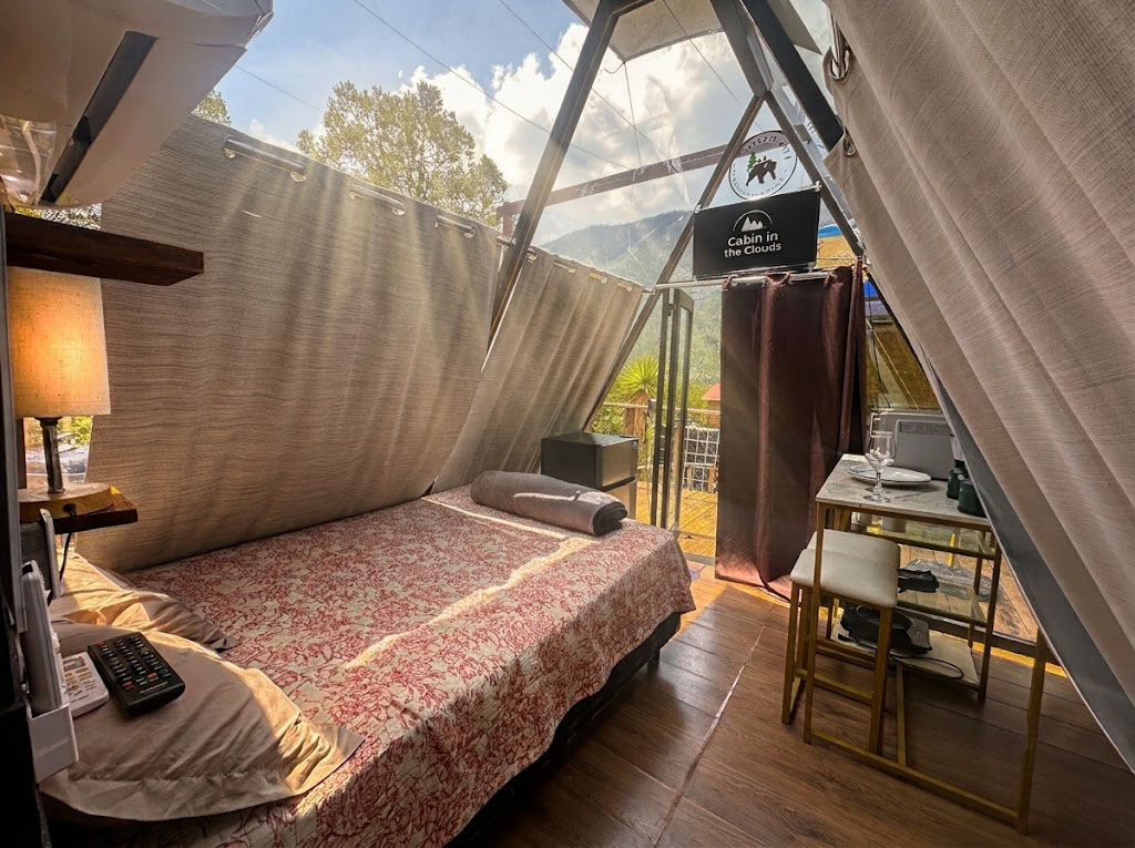 Recámara acogedora de SIRIUS con cama vestida, lámpara cálida y vista panorámica a la sierra a través del ventanal de cristal