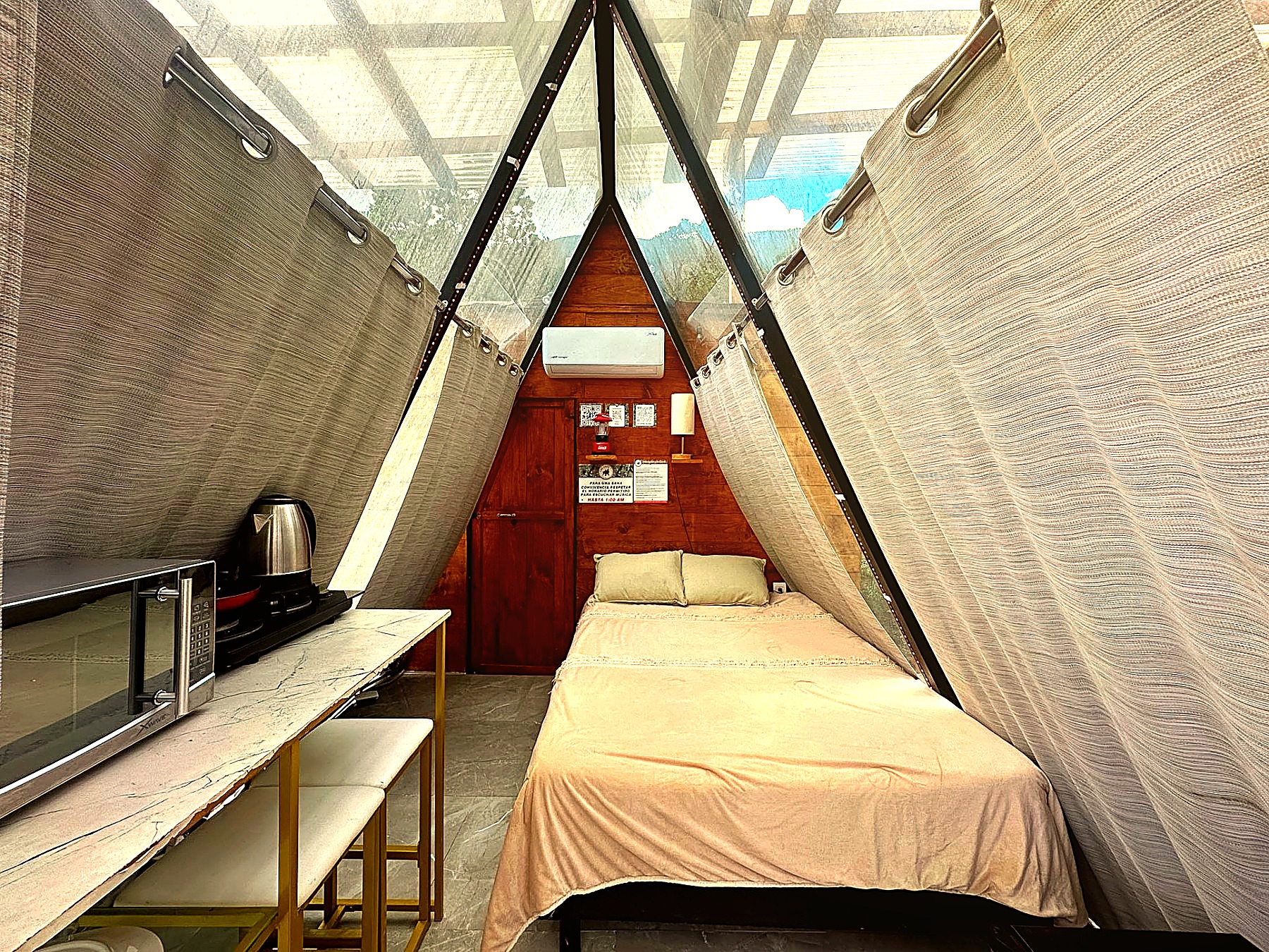 Interior de ATENEA con cama matrimonial, cocineta equipada, climatización y cortinas texturizadas bajo el ventanal A-frame