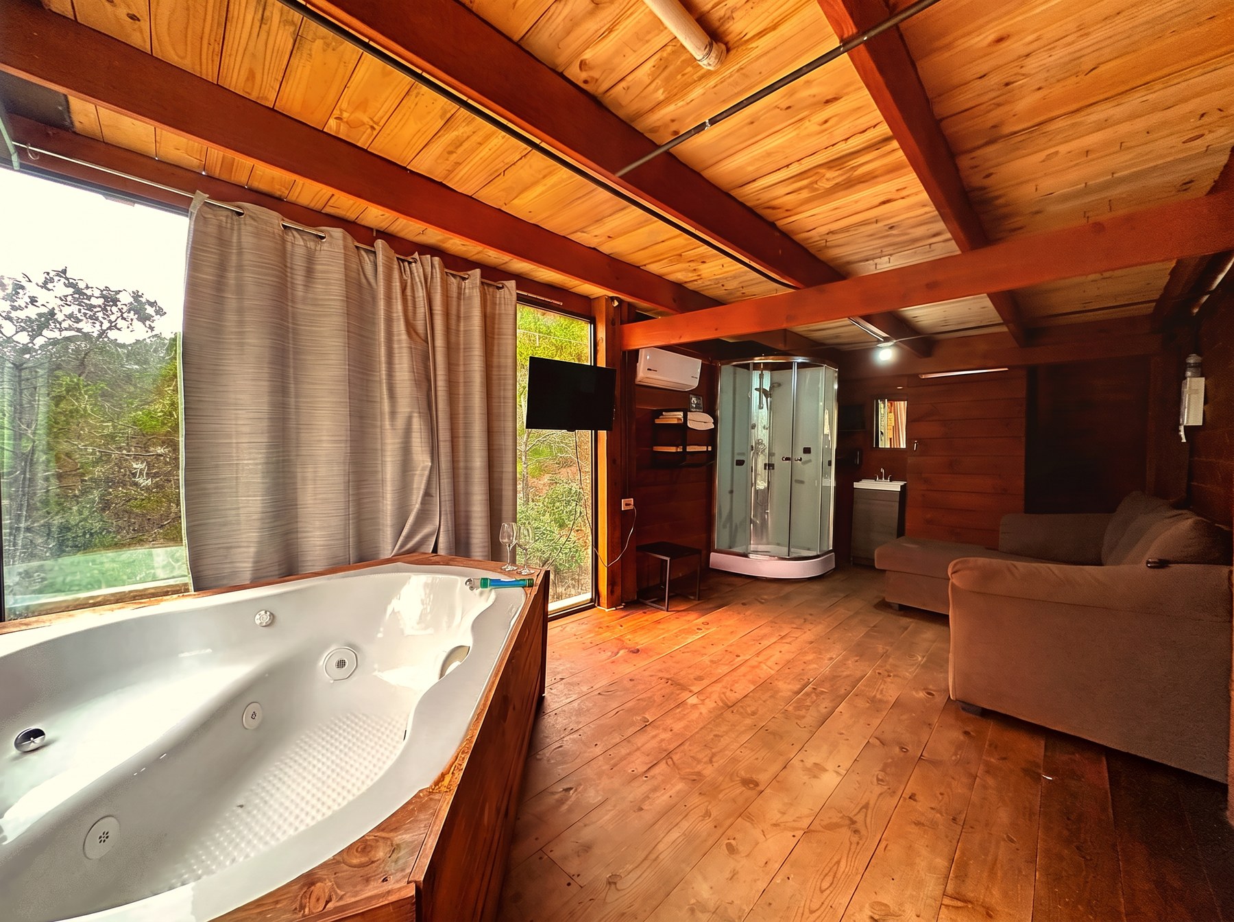Jacuzzi privado de ATENEA junto a sala de estar con sillón, regadera con hidromasaje y ventanal al bosque